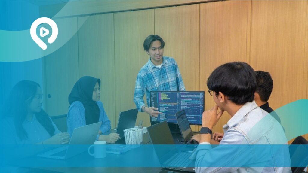 Tim pengembang website sedang berdiskusi dan bekerja menggunakan laptop serta komputer dalam sebuah ruang kerja.
