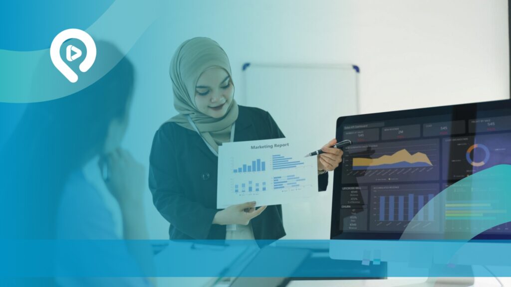 Perempuan berhijab sedang mempresentasikan laporan pemasaran sambil menganalisis data pada dashboard komputer.